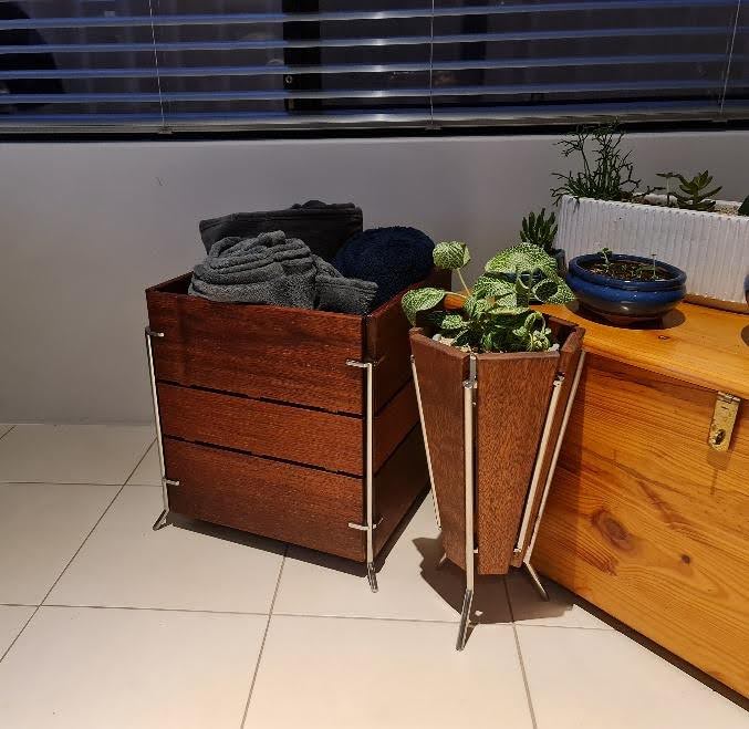 Two wooden storage containers with metal frame supports placed on a tiled floor indoors. The larger container on the left holds neatly folded dark towels, while the smaller container on the right holds green potted plants. A wooden chest and additional potted plants sit in the background near a window with horizontal blinds.